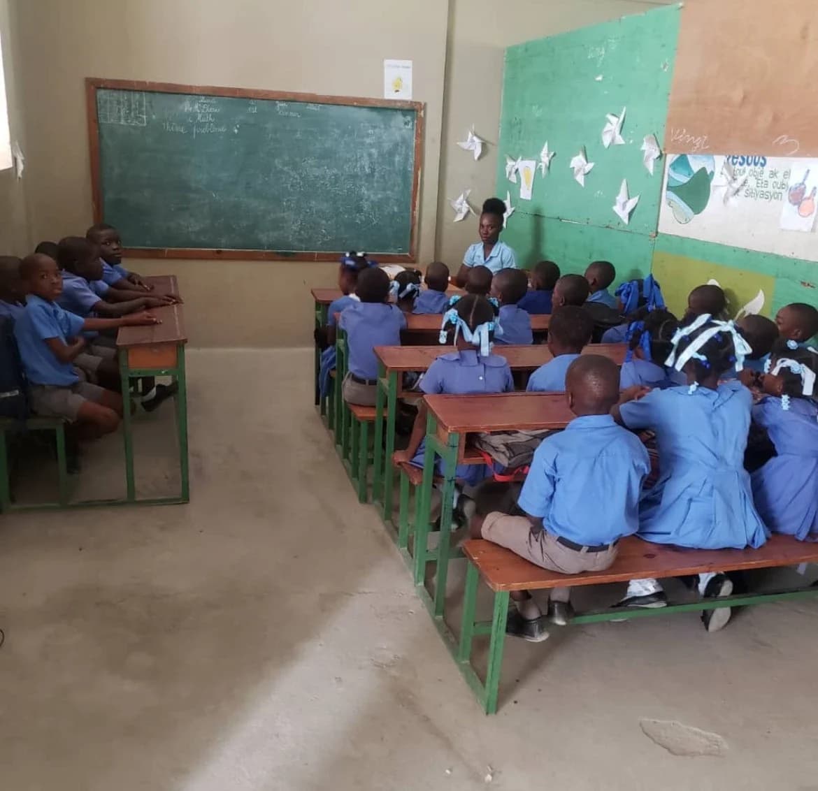 Students in blue uniforms sit at wooden desks facing their teacher