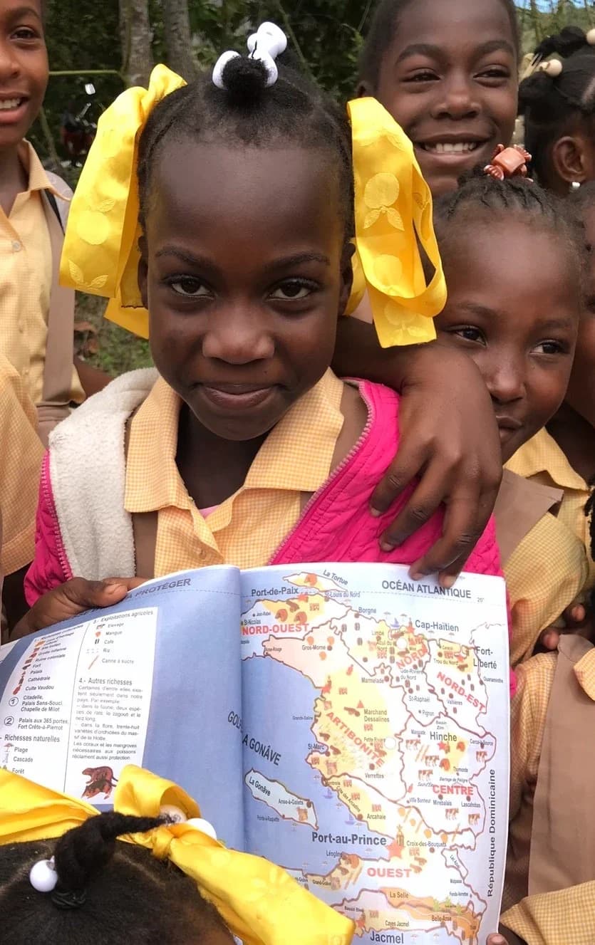A smiling girl in a yellow uniform holds up a map of Haiti in her schoolbook