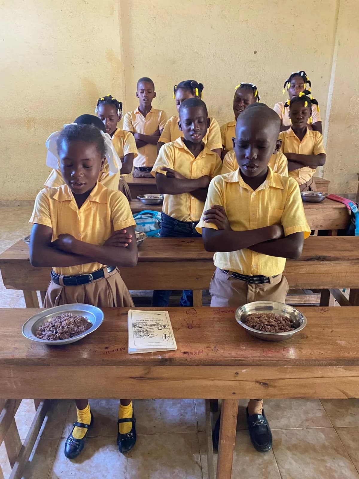 Students in yellow uniforms pray over their meal, a schoolbook on the desk between their bowls