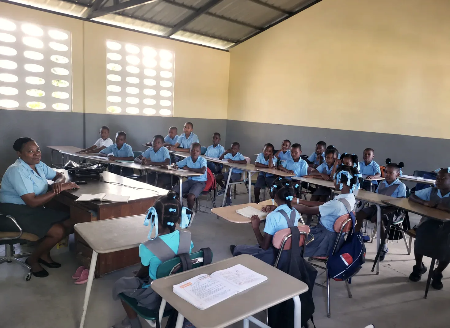 A teacher sits at her desk before a classroom of students in light-blue uniforms