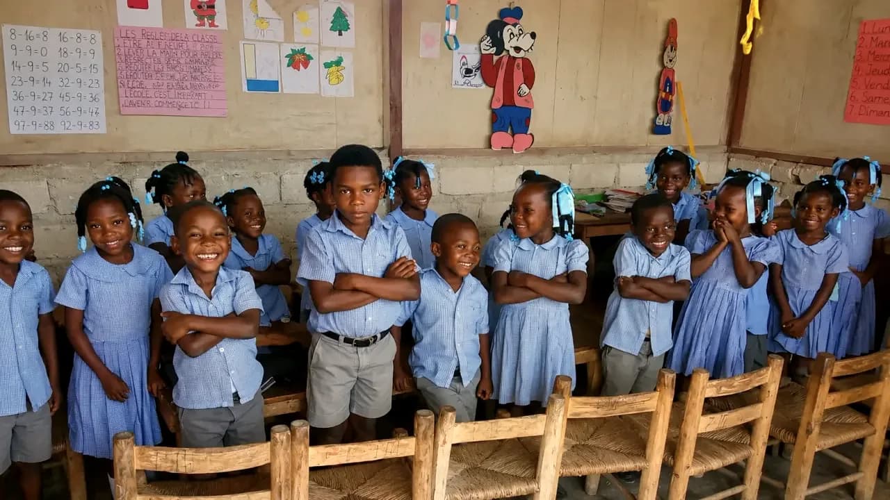 Smiling young students in blue uniforms stand beside their wooden chairs in an HEF classroom